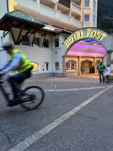 Ein Radfahrer in einer gelben Weste fährt in der Abenddämmerung am Eingang des Hotels Post vorbei und gleitet über die Strecke, während zwei Personen in der Nähe von Metallbarrieren vor dem beleuchteten Hotel stehen.