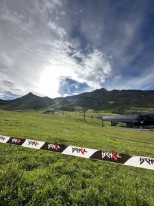 Ein grasbewachsener Hang mit Skiliftanlagen unter leicht bewölktem Himmel und strahlender Sonne. Ein schwarz-weißes Band mit roten Logos grenzt einen Teil der grünen Wiese ab und deutet auf eine Mountainbike-Strecke hin. Im Hintergrund erheben sich Berggipfel.