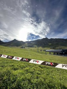 Ein grasbewachsener Hang mit Skiliftanlagen unter einem bewölkten Himmel, die Sonne scheint hinter den Bergen in der Ferne und eine Barriere mit der Aufschrift „TIGNES“ im Vordergrund markiert den Beginn eines aufregenden MTB-Trails.