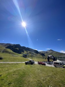 Eine helle Sonne scheint über eine grasbewachsene Berglandschaft mit ein paar geparkten Autos, einem kleinen Gebäude und Hügeln im Hintergrund unter einem klaren blauen Himmel – perfekt, um die nahegelegene Mountainbike-Strecke zu genießen und sich der Mountainbike-Bewegung hinzugeben.