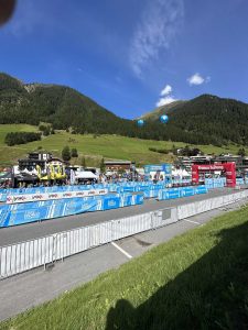 Ein spannendes Mountainbike-Rennen in einem malerischen Tal – Absperrungen, blaue Sponsorenbanner, Zelte und Zuschauer säumen die Strecke; grasbewachsene Hügel, Bäume und ein blauer Himmel mit Wolken und Ballons runden die lebendige Szene ab.