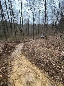 An einem bewölkten Tag schlängelt sich ein Feldweg durch einen Wald mit kahlen Bäumen. Rechts vom Weg steht ein kleiner Bagger, umgeben von Laub – ein Hinweis auf die jüngste Mountainbike-Bewegung in der Gegend.
