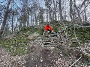 Eine Person in einer leuchtend roten Jacke hockt auf einer moosbewachsenen Steinmauer neben einer Mountainbike-Strecke in einem Wald mit kahlen Bäumen und abgefallenem Laub. Der Himmel ist bedeckt, was der kühlen, winterlichen Szenerie eine abenteuerliche Mountainbike-Atmosphäre verleiht.