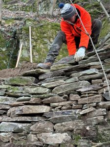 Eine Person in orangefarbener Jacke und Handschuhen repariert eine Steinmauer im Freien entlang einer Mountainbike-Strecke und platziert dabei sorgfältig Steine. Im Hintergrund sind Gartengeräte und Grünpflanzen zu sehen, die den Geist der Mountainbike-Bewegung verkörpern.