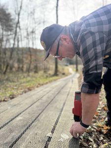 Eine Person mit Mütze und Brille tackert Drahtgeflecht auf eine Mountainbike-Strecke entlang eines Holzwegs in einem Waldgebiet. Auf dem Boden liegen verstreute Blätter, im Hintergrund sind Bäume zu sehen.