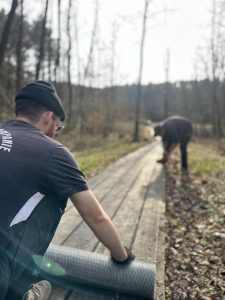Zwei Personen rollen gemeinsam eine große Matte entlang einer hölzernen Mountainbike-Strecke in einem Waldgebiet aus. Eine Person steht nah an der Kamera, die andere in einiger Entfernung. Beide konzentrieren sich auf die Aufgabe.