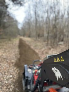 Ein verschwommenes Bild eines Feldwegs, der in einem Wald geräumt wird, mit einer Nahaufnahme von Baumaschinen mit einem Wolfslogo und Sicherheitswarnsymbolen – perfekt für das nächste Mountainbike-Abenteuer.