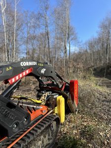 Eine Nahaufnahme eines Cormidi-Ketten-Minikippers mit Anbaugeräten auf einem Mountainbike-Trail, umgeben von blattlosen Bäumen unter einem klaren blauen Himmel.