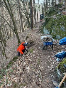 Eine Person in einer orangefarbenen Jacke hockt an einem Feldweg im Wald und legt Steine für den Bau einer Mountainbike-Strecke auf. Eine Schubkarre voller Steine, eine Schaufel und eine blaue Tasche stehen in der Nähe. Bäume und moosbedeckte Felsen umgeben die Szene.