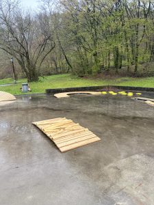Ein Außenbereich aus nassem Beton mit als Laufstrecke angeordneten Holzplanken, gelben Trittflächen und einer Mountainbike-Atmosphäre vor der Kulisse grüner Bäume und Gras an einem regnerischen Tag.