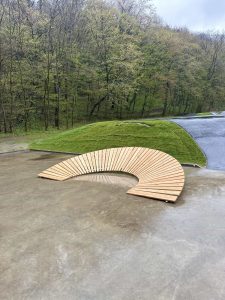 Eine geschwungene Holzbank steht im Freien auf einer nassen Betonoberfläche neben einem Pfad mit grünen Grashügeln und einem Wald aus Bäumen im Hintergrund.