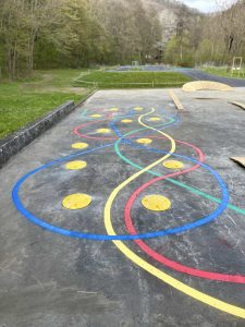 Bunte, gewundene Linien in Blau, Gelb und Rot sind auf eine Mountainbike-Strecke aus Beton im Freien gemalt und kreuzen sich mit runden gelben Markierungen. Der Weg ist von Gras und Bäumen in einer parkähnlichen Umgebung umgeben.