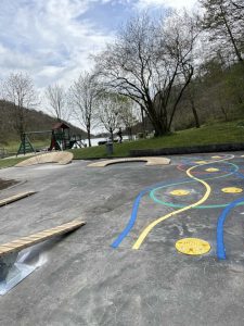 Ein Spielplatz mit bunten Linien und Kreisen auf dem Beton, Seilen zum Balancieren, einer Schaukel, Bäumen und einem See im Hintergrund – und in der Nähe ein kleiner Mountainbike-Track unter einem teilweise bewölkten Himmel.