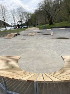 Eine geschwungene Holzbank im Vordergrund umgibt eine Betonfläche mit Mountainbike-Rampen und Streckenkonstruktionen, im Hintergrund sind unter einem bewölkten Himmel Bäume, Gras und ein Spielplatz mit Schaukeln zu sehen.
