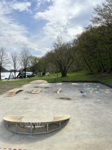 Ein Skatepark aus Beton mit geschwungenen Bänken, Rampen und bunten aufgemalten Mustern liegt neben einer grasbewachsenen Parkfläche mit Bäumen, einem See und einem nahe gelegenen Mountainbike-Trail unter einem teilweise bewölkten Himmel.