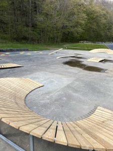 Gebogene Holzbänke stehen auf einer Betonfläche mit Pfützen, umgeben von grünen Bäumen und einer Rasenfläche in der Nähe eines Mountainbike-Trails. Im Hintergrund sind farbige Kreise auf den Boden gemalt.