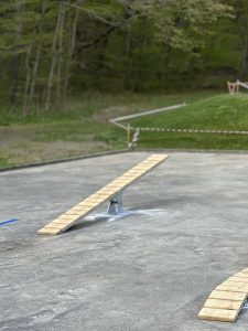 Eine Holzwippe mit Metallsockel steht auf einer Betonfläche im Freien, in der Nähe eines Mountainbike-Trails. Im Hintergrund sind Gras, Bäume und Absperrband zu sehen.