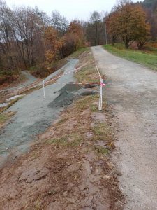 Ein von Gras und Bäumen gesäumter Schotterweg schlängelt sich durch einen Park. An den Stellen, wo der Boden aufgerissen wurde – möglicherweise als Vorbereitung für zukünftige Mountainbike- oder MTB-Strecken unter einem bewölkten Himmel – ist er mit rot-weißem Absperrband markiert.