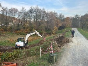Ein kleiner Bagger gräbt einen Graben entlang einer unbefestigten Straße und schafft so etwas wie eine Mountainbike-Strecke. Zwei Personen stehen daneben; eine deutet in die Ferne. Das Gebiet ist von herbstlich gefärbten Bäumen umgeben und mit rot-weißem Band abgesperrt.