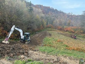 Zwei Bagger graben und bewegen Erde auf einem grasbewachsenen, unebenen Feld und formen so einen neuen Mountainbike-Trail, gesäumt von herbstlich gefärbten Bäumen. Im Hintergrund erhebt sich ein bewaldeter Hang unter einem leicht bewölkten Himmel.