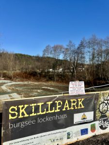 Ein Schild des Skillpark Burgsee Lockenhaus steht vor einer Mountainbike-Strecke, umgeben von Bäumen, Hügeln und strahlend blauem Himmel. Die Gegend fängt den Geist der Natur und der Mountainbike-Bewegung in diesem malerischen Outdoor-Park ein.