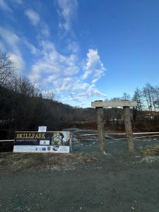 Neben zwei aufrecht stehenden Betonpfeilern auf einer Schotterfläche steht ein Schild mit der Aufschrift „Skillpark“ und weist auf nahegelegene Mountainbike-Strecken inmitten kahler Bäume und eines teilweise bewölkten blauen Himmels hin.