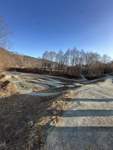 Ein Dirtbike-Trail mit gewundenen Pfaden und Unebenheiten ist von blattlosen Bäumen unter einem klaren blauen Himmel umgeben, die im Nachmittagssonnenlicht lange Schatten werfen – eine perfekte Szene für Mountainbike-Enthusiasten und die Mountainbike-Bewegung.