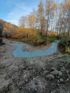 Ein frisch gegossener Betonweg schlängelt sich durch eine schlammige, ländliche Landschaft mit kahlen Bäumen und Herbstlaub auf beiden Seiten und lädt zum Mountainbiken unter einem klaren blauen Himmel ein.