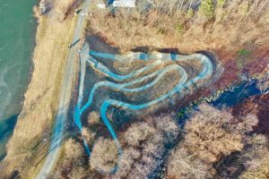 Luftaufnahme einer kurvenreichen Mountainbike-Strecke mit blauen Linien, die durch ein Waldgebiet neben einem Gewässer und einer unbefestigten Straße führt. Entlang des Weges sind vereinzelte Bäume und blattlose Äste zu sehen.