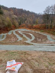 Ein geschwungener Schotterweg schlängelt sich durch ein Stück nackten Bodens, umgeben von herbstlichen Bäumen. Im Vordergrund liegt neben dem Weg eine weiß-orangefarbene Tasche. Im Hintergrund erheben sich bewaldete Hügel unter einem bewölkten Himmel.