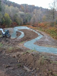 Ein Projekt zur Renaturierung eines gewundenen Baches in einem Waldgebiet mit frisch geformter Erde, die eine mögliche Mountainbike-Strecke bildet, und einem kleinen Bagger auf der linken Seite; Bäume mit Herbstlaub umgeben die Szene.