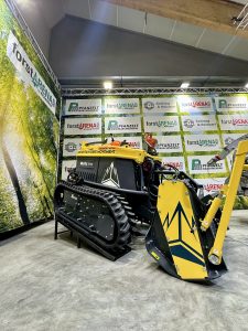 Eine kompakte, gelbe Raupen-Forstmaschine mit schwarz-weißer Gebirgsgrafik steht in einem Ausstellungsraum mit Bannern zum Thema Wanderwege und Firmenlogos an den Wänden.