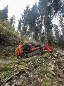 Eine kompakte orange-schwarze Forstmaschine mit Raupenfahrwerk steht an einem felsigen, moosbedeckten Hang neben einem gewundenen MTB-Pfad in einem Wald mit hohen immergrünen Bäumen unter einem bedeckten Himmel.